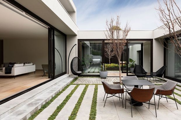 A contemporary private courtyard enclosed by white rendered walls and large dark-framed glazed openings. Linear stone paving is interspersed with strips of grass, leading to a central planted area with slender trees. Outdoor dining furniture and lounge chairs occupy the courtyard, while a living room with pale timber flooring and a white sofa is visible through the open glazing. The design is clean-lined, minimalist, and strongly connected to the indoor and outdoor spaces.