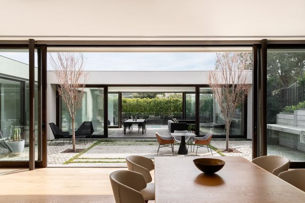 A refined contemporary dining area opens through expansive dark-framed glazing onto a landscaped courtyard. A light timber dining table and sculptural bowl occupy the foreground, while an outdoor seating area, tiled paving, slender trees, layered glazing, and raised concrete planters create a calm indoor–outdoor connection. The composition is bright, spacious, and minimalist with strong horizontal lines.
