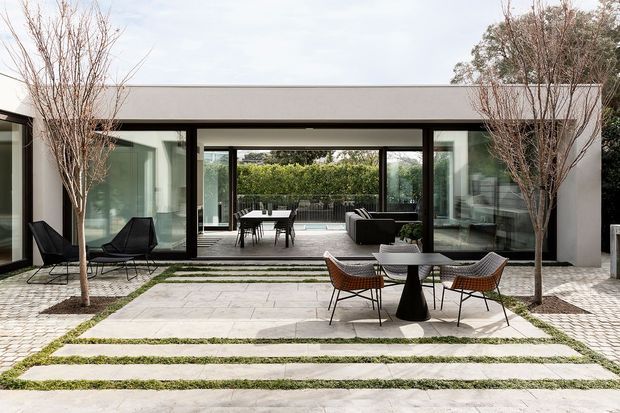 Contemporary single-storey courtyard house viewed from the landscaped outdoor area. Large floor-to-ceiling glazed openings connect the paved courtyard to an interior living space and a dining area beyond. The restrained white and dark-grey building envelope is framed by slim dark window and door frames, while broad pale stone paving is intersected by narrow strips of grass. Outdoor lounge and dining furniture occupy the foreground, with additional seating visible inside.
