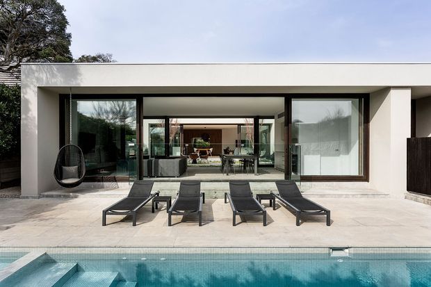 Contemporary single-storey poolside house with a broad flat roof, white rendered facade, dark-framed floor-to-ceiling glazing, and an open connection between the interior living area and paved terrace. Four sun loungers and a hanging chair sit beside a turquoise swimming pool, with trees visible beyond.