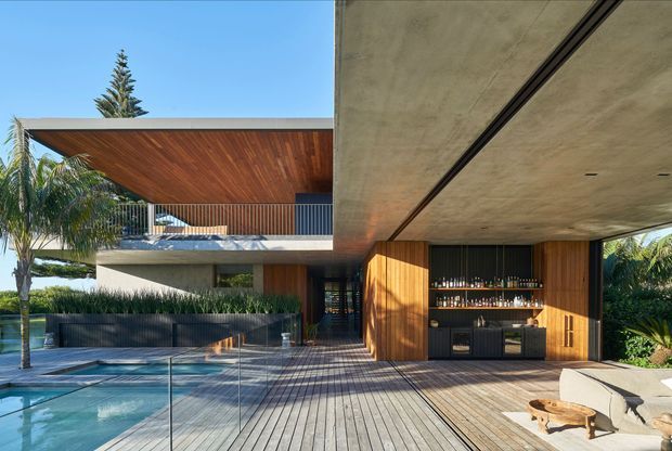 Contemporary multi-level residence viewed from a poolside timber deck, featuring strong cantilevered concrete forms, warm timber soffits and wall cladding, extensive glazing, a glass pool barrier, and an integrated outdoor bar beneath a deep overhang. Tropical planting and bright daylight create a resort-like outdoor setting.