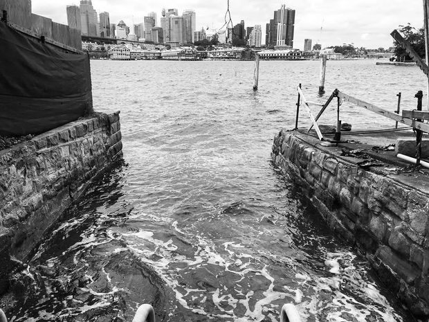 Black-and-white photograph looking out from a narrow stone-lined waterfront channel or slipway toward a broad body of water. Rough masonry retaining walls, mooring posts, ropes, and dock infrastructure frame the foreground, with an urban skyline and waterfront buildings across the water.