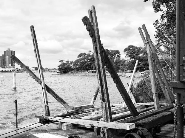Black-and-white photograph of a weathered timber waterside structure or working dock, with rough posts, beams, ropes, and a partially visible corrugated or woven-sided enclosure in the foreground. A broad body of water, tree-lined shoreline, and distant buildings are visible beyond. The image has a documentary, rustic character and strong monochrome contrast.