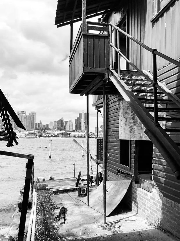 Black-and-white exterior photograph of a weathered waterside building with corrugated cladding, dark timber boards, exposed brickwork, and prominent external metal stairs and balconies. The structure sits directly beside a waterfront with a city skyline, marina, and piers visible across the water. The scene has an industrial, utilitarian character and strong vertical and diagonal structural lines.