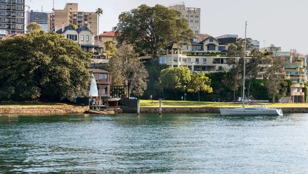 Waterfront view across a broad body of water toward a landscaped urban residential shoreline. Mature trees, a grassy foreshore, small boats and marina-like moorings sit in front of a mix of older character houses, apartment buildings and newer multi-storey developments. The image is an exterior urban waterfront scene dominated by the water and shoreline rather than a single building.