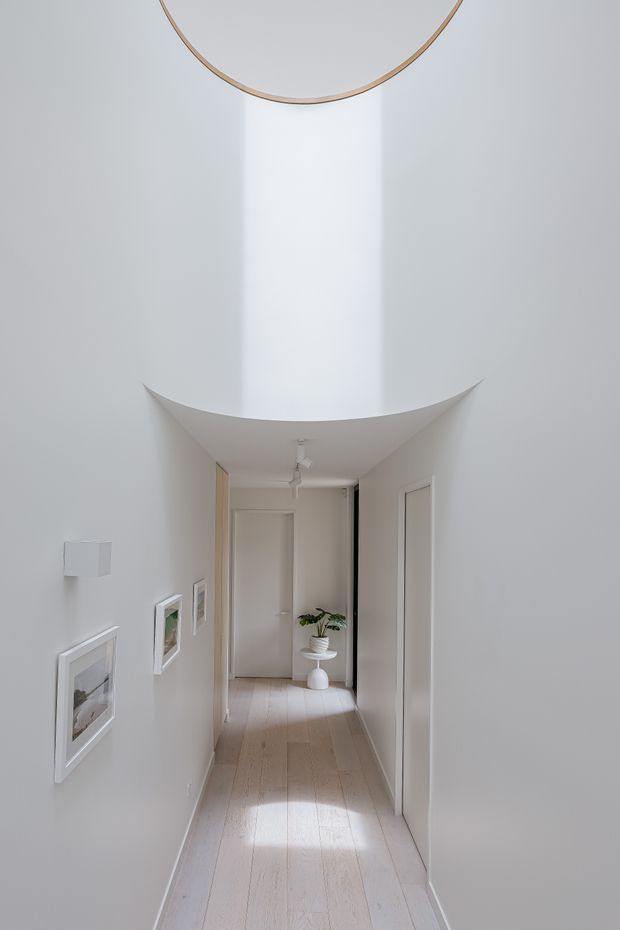 A bright, minimalist residential hallway with white walls and doors, pale timber flooring, framed artwork, and a small potted plant at the far end. The corridor features a distinctive curved ceiling transition or sculptural opening that creates a dramatic vertical void above the passage.