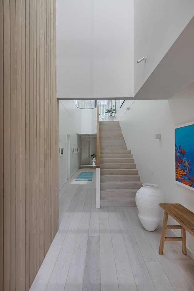 Bright minimalist interior hallway with a central open staircase. Pale whitewashed timber flooring, white walls, and vertical light-wood wall panelling create a calm, cohesive palette. A large white sculptural vessel, simple timber bench, colourful artwork, and glimpses of an upper landing and pendant light complete the space.