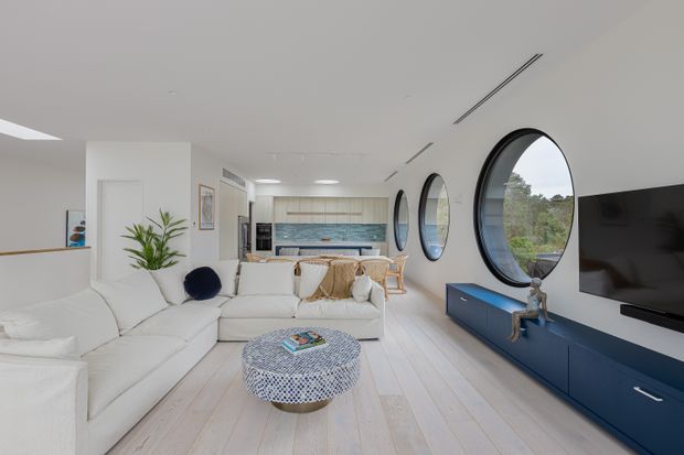 Bright open-plan living room with a large white sectional sofa, patterned round coffee table, navy built-in media cabinetry, wall-mounted television, and a compact dining and kitchen area beyond. The room features pale timber flooring, white walls and ceiling, large circular black-framed windows, and blue-toned kitchen cabinetry with a tiled backsplash.
