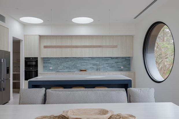 Contemporary kitchen and dining area with pale wood cabinetry, a long dark island with light benchtop, blue-grey stacked-stone splashback, integrated appliances, and a large oval window. A dining table with upholstered seating occupies the foreground.