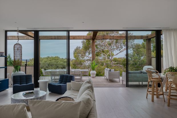 A bright, open-plan living room with a large cream sectional sofa, dark lounge chairs, a textured round coffee table, and a dining area with rattan chairs. Floor-to-ceiling black-framed glazing opens onto a timber deck and covered outdoor lounge, with leafy views beyond. The interior has light timber flooring, neutral furnishings, abundant natural light, and a relaxed indoor-outdoor connection.