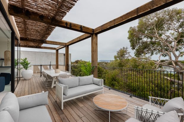 A spacious elevated outdoor terrace with timber decking, white outdoor sofas and lounge chairs, a round timber coffee table, black metal balustrades, and a timber pergola with a woven overhead screen. The terrace overlooks a leafy suburban landscape and is connected to the interior through large glazed doors.