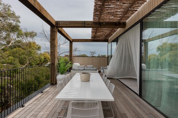 A spacious elevated outdoor dining terrace with a long white dining table and woven chairs, adjoining a contemporary glazed interior. Timber decking, substantial timber pergola beams, and a woven reed canopy create a warm natural setting overlooking bushland. Large sliding glass doors with sheer curtains connect the terrace to the living space.