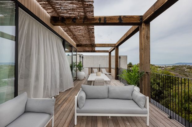A contemporary elevated outdoor terrace with timber decking, a light upholstered sofa and armchair, a dining table, and a timber pergola covered with woven shade screening. Full-height glazing with sheer curtains connects the terrace to the interior, while dark metal balustrades frame views across the coastal landscape.