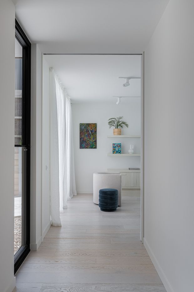 A bright, minimalist interior view through a hallway into a compact home office or study. White walls and ceiling, pale timber flooring, sheer full-height curtains, black-framed glazing, recessed shelving, a light built-in desk, a white upholstered chair, and a dark blue rounded ottoman create a clean, calm atmosphere. A colourful abstract artwork and potted plant provide restrained accents.