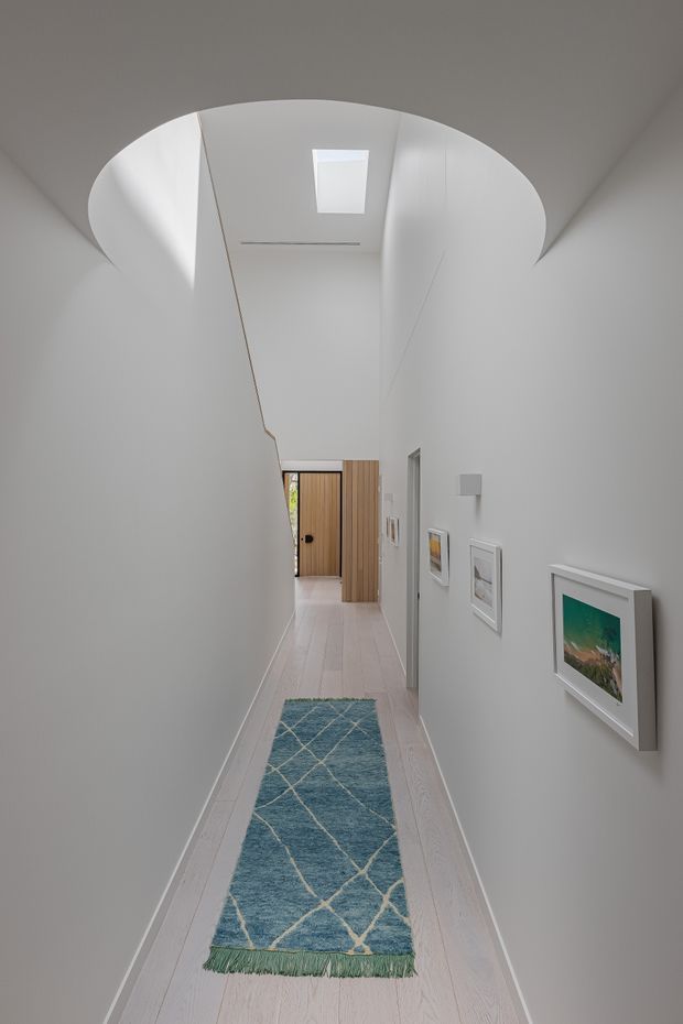 A narrow, bright residential hallway with white walls and pale timber flooring. A long blue patterned runner leads toward a timber front door, with framed artwork along the right wall. The ceiling features a large curved opening or sculptural recessed form with a skylight, creating a distinctive architectural focal point.