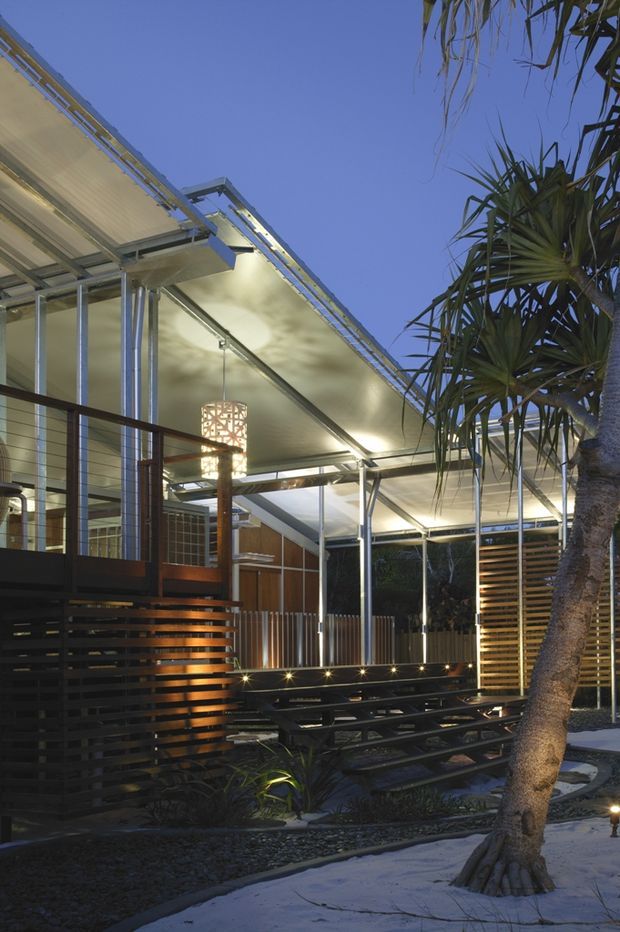 A dusk exterior view of a contemporary pavilion or covered outdoor entertaining area. The structure features a lightweight translucent roof with exposed slender metal framing, extensive glazing, timber-toned horizontal privacy screens, and a large decorative pendant light. Tiered built-in seating and low landscape lighting occupy the landscaped courtyard, with tropical-style planting and a mature tree in the foreground.