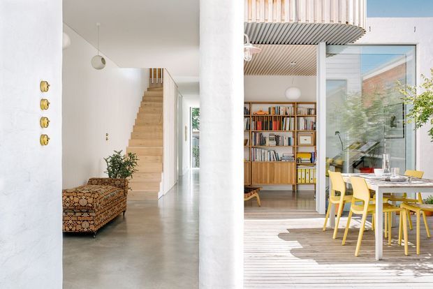 Bright, open-plan residential interior showing a white plastered entry hall with a light timber staircase, concrete-look floor, and patterned upholstered bench. Through the opening is a timber-lined study or living area with full-height shelving and books, opening onto a sunlit timber deck with a white dining table and yellow chairs. The space has abundant glazing, a high ceiling, and a clean contemporary feel.