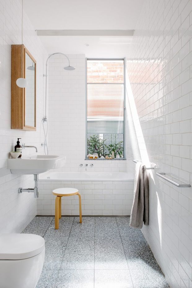 Bright, narrow bathroom with white subway-tiled walls, a freestanding bathtub beneath a tall window, wall-mounted white basin, exposed plumbing, and a walk-in shower with twin showerheads. A timber-framed mirrored cabinet, terrazzo-style grey floor tiles, simple towel rail, and a small yellow stool add warmth and contrast. The space has a clean, airy contemporary character with abundant natural light.