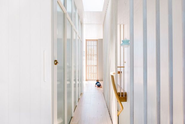 A bright, narrow residential hallway with white vertical wall panelling, pale timber flooring, extensive internal glazing and framed glass doors, and a timber stair handrail visible on the right. A slatted timber screen and skylight enhance the airy linear space.