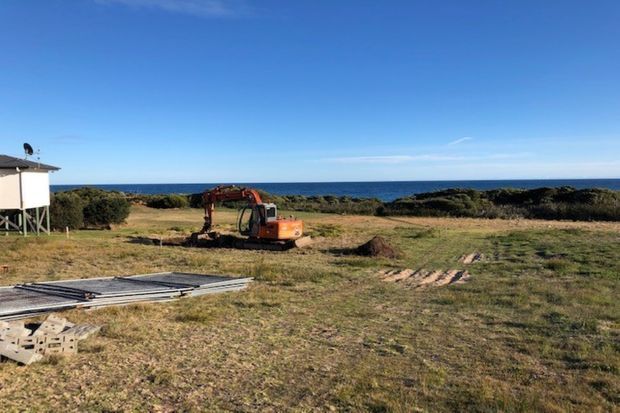 A coastal vacant building site viewed toward the ocean, with an excavator working on the grassy terrain, construction materials in the foreground, and a small elevated structure at the left edge. The image is a straightforward outdoor site/construction view under clear blue sky.