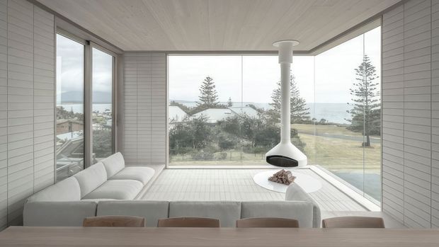Bright, minimalist coastal living and dining space enclosed by expansive floor-to-ceiling glazing, with pale tiled wall and floor surfaces, a low light-coloured sectional sofa, a suspended white fireplace, and a long timber dining table. The windows overlook a seaside residential landscape.