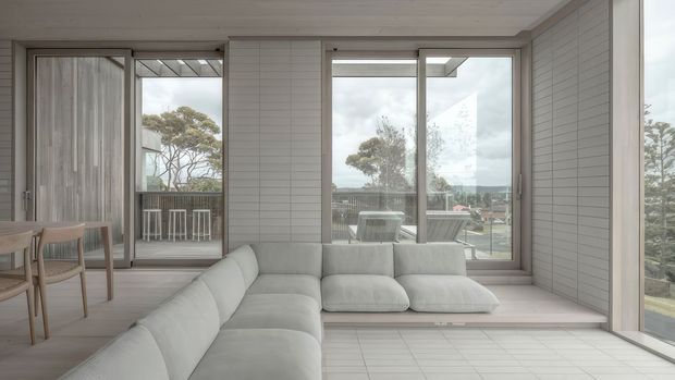 Bright, airy contemporary living room with a large pale sectional sofa, white horizontal wall panelling, light tiled flooring, and extensive floor-to-ceiling glazing. Sliding glass doors open to a balcony with outdoor bar seating, while trees and a coastal-looking landscape are visible beyond. The space has a restrained, monochromatic palette and strong indoor-outdoor connection.