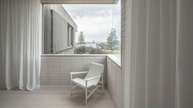 A quiet, minimalist enclosed balcony or sunroom with floor-to-ceiling glazing, sheer white curtains, pale timber flooring, and light rectangular wall tiling. A single white lounge chair faces the view, while a neighbouring modern building, trees, and water are visible beyond the tiled parapet.