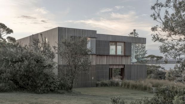 A contemporary two-storey house viewed from a landscaped garden, composed of dark vertical timber cladding with a strong rectilinear, flat-roofed form. Large windows and a recessed ground-floor opening are partially screened by mature shrubs and ornamental grasses. A distant body of water is visible beyond the property, contributing to a quiet coastal or waterfront setting.
