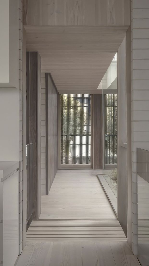 A narrow, light-filled interior passage with pale timber flooring, walls and overhead soffit. Vertical timber screening encloses glazed openings at the far end, while a skylight or clerestory opening brings in daylight from above. White horizontal wall cladding and dark timber door panels add contrast, creating a calm, minimalist architectural corridor with a strong focus on linear geometry and filtered light.