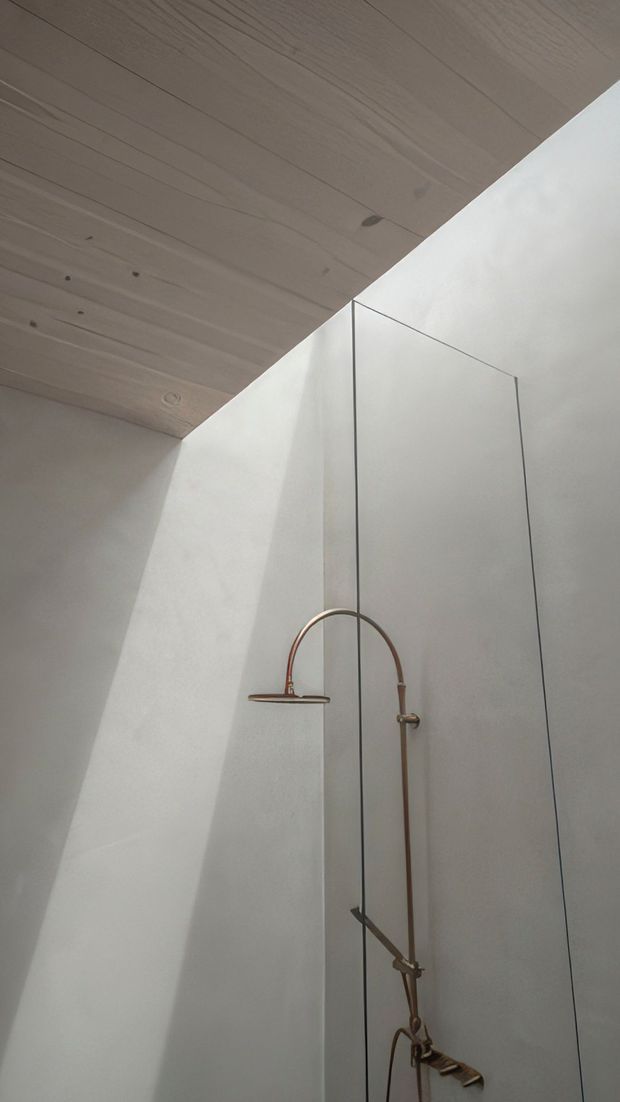 Minimalist shower area with a frameless glass shower screen, white plastered walls, and a pale timber-lined sloping ceiling. A copper-toned exposed shower pipe and fixtures provide the main decorative detail, with soft natural light creating a calm, understated atmosphere.