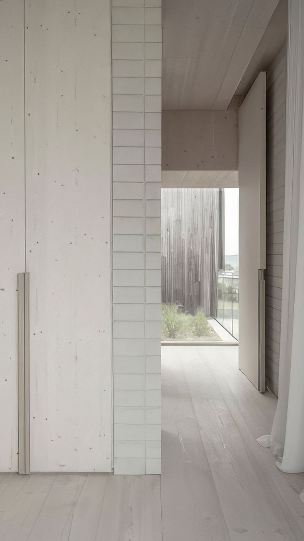 A narrow, minimalist interior passage with pale timber-look flooring, white tiled and plasterboard walls, and a light timber or painted flush door. The corridor opens toward an exterior courtyard or terrace with vertical weathered cladding, planting, and a railing visible beyond. The palette is very light and restrained, with clean linear detailing and strong natural daylight.