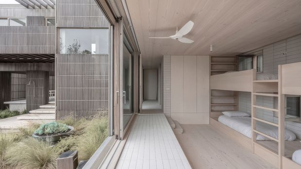 A bright, compact contemporary bedroom or sleeping area opening onto a planted courtyard. The room features pale timber flooring and ceiling boards, full-height sliding glazing, built-in bunk-style beds and shelving, and a minimal white ceiling fan. The courtyard uses vertical timber screening, paving, ornamental grasses, and low planting, creating a calm indoor-outdoor connection.