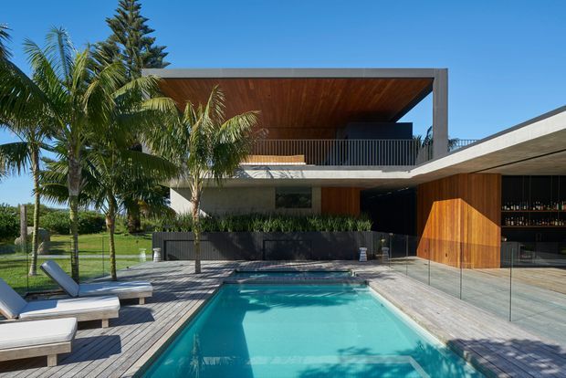 A contemporary two-storey residence with strong indoor-outdoor connection, featuring a turquoise swimming pool, timber pool deck, glass balustrades, lush tropical palms, dark masonry planter walls, and extensive warm timber soffits and cladding. The upper level has a deep covered balcony with a large rectangular roof opening, while the ground floor includes a glazed entertaining area and a timber-clad feature door.