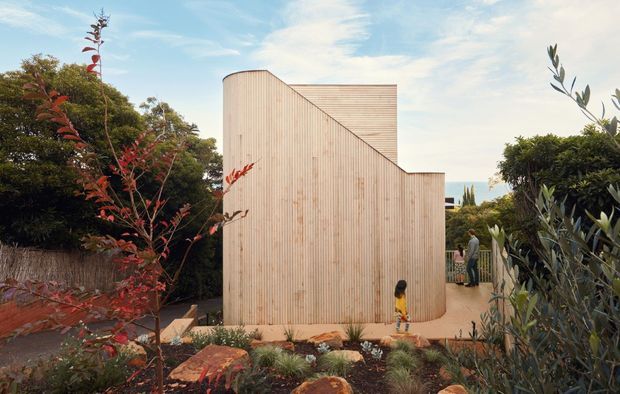Exterior view of a sculptural, single-storey building clad in pale vertical timber boards. The facade has a curved corner and a distinctive stepped, sloping roofline, set within a landscaped garden with paved paths and mature planting. A glimpse of blue water or sky is visible beyond the property.
