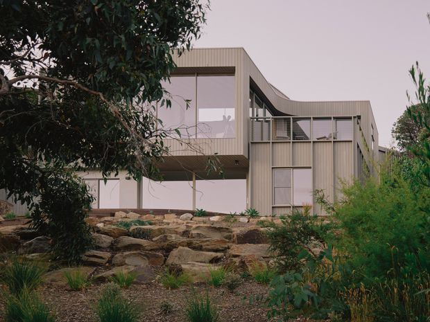 Exterior view of a contemporary multi-level house set into a landscaped rocky garden. The building features light vertical ribbed cladding, large glazed openings, projecting upper volumes, and a gently curved roofline or facade edge. Native-style planting and exposed rock landscaping surround the structure.
