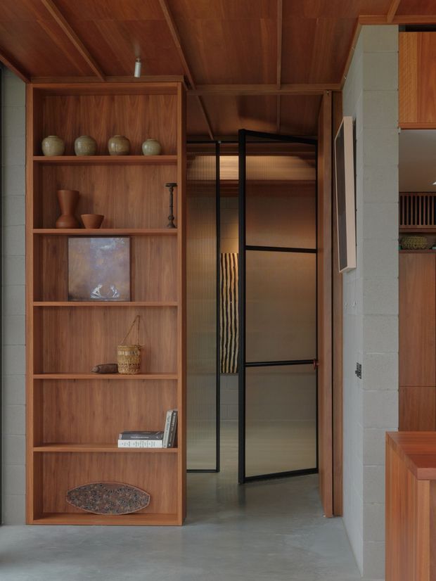 A warm, carefully detailed interior passage featuring a full-height built-in timber shelving unit displaying ceramics, artwork, books, and decorative objects. Black-framed translucent glass doors open into an adjacent space, while the surrounding timber-lined ceiling and exposed painted masonry wall contribute to a refined modern interior with strong natural material character. A polished concrete floor and dark metal door framing provide contrast.