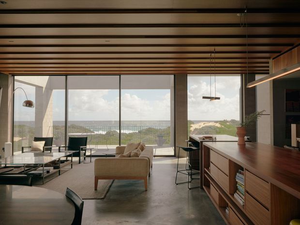 A refined open-plan living, dining and kitchen interior with expansive floor-to-ceiling glazing overlooking a coastal landscape. The room features a warm timber slatted ceiling, exposed concrete flooring, extensive timber built-in cabinetry, a kitchen island, contemporary lounge seating, a glass coffee table, and suspended linear pendant lighting. The composition is warm and sophisticated, with strong indoor–outdoor connection and ocean views.