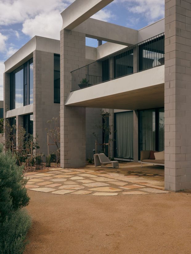 A contemporary multi-storey residential courtyard viewed from outdoors, featuring pale masonry block walls, strong rectangular concrete-like structural frames, extensive floor-to-ceiling glazing, dark metal vertical balustrades, and sheltered outdoor living areas. The courtyard is paved with irregular warm-toned stone slabs and includes lounge seating and sparse landscaping. The architecture has a sculptural, geometric composition with open-air terraces and a partly visible blue sky.