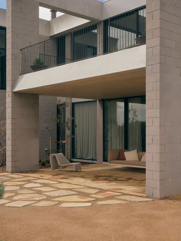 A contemporary multi-level courtyard facade with pale masonry block walls, deep concrete overhangs, dark-framed glazing, and black vertical metal balcony railings. The ground-level covered outdoor lounge contains modern seating and potted plants, opening onto an irregular natural-stone paved courtyard and muted gravel or rendered ground plane. The composition emphasizes strong geometric forms, recessed glazing, and a sheltered outdoor living area.
