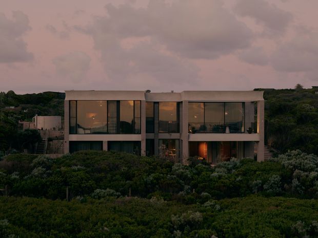 A dusk exterior photograph of a large contemporary two-storey residence set within dense natural landscaping. The home has a restrained rectangular form, pale rendered or concrete-like facade surfaces, extensive floor-to-ceiling glazing, recessed balconies or terraces, and warm interior lighting visible through the windows. The surrounding vegetation and darkening sky create a moody, secluded atmosphere.