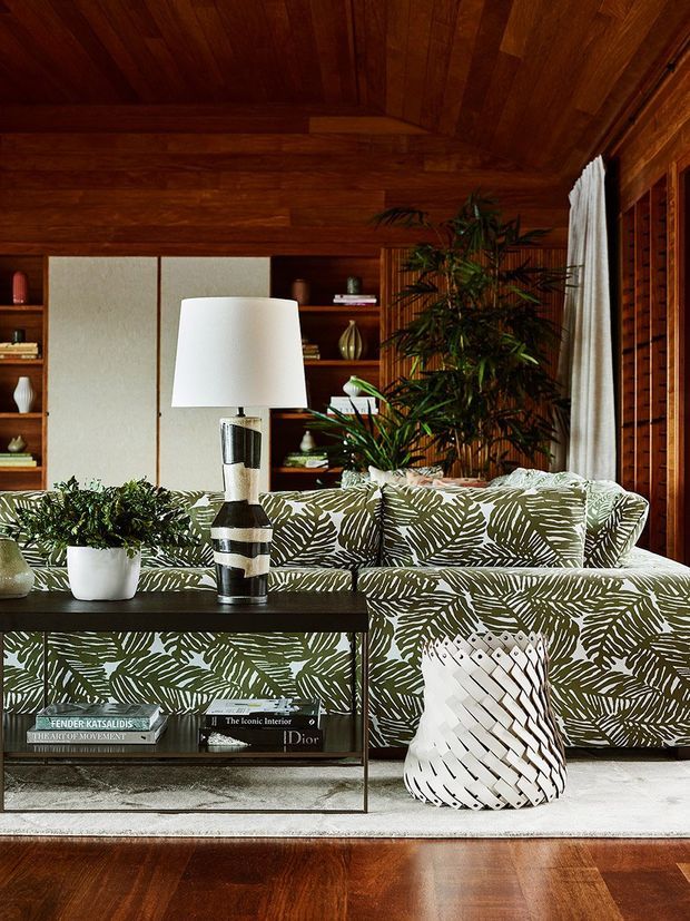 A richly layered living room with warm timber walls and a slatted timber ceiling, built-in shelving, and a large patterned sectional sofa in botanical green and cream upholstery. A dark open console table, sculptural table lamp, potted plants, woven white planter, and neutral rug complete the refined, nature-inspired interior.