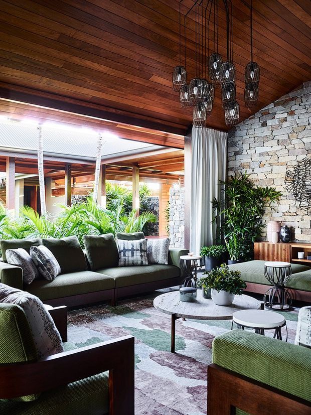 A richly layered living room with a warm timber-lined ceiling, exposed pale stone feature wall, and large openings to a lush planted courtyard. The room contains a deep green sectional sofa, matching lounge chairs, patterned cushions, round marble or stone coffee tables, indoor plants, and a dramatic cluster of suspended wire pendant lights. The overall composition blends indoor and outdoor living with abundant natural light and earthy materials.