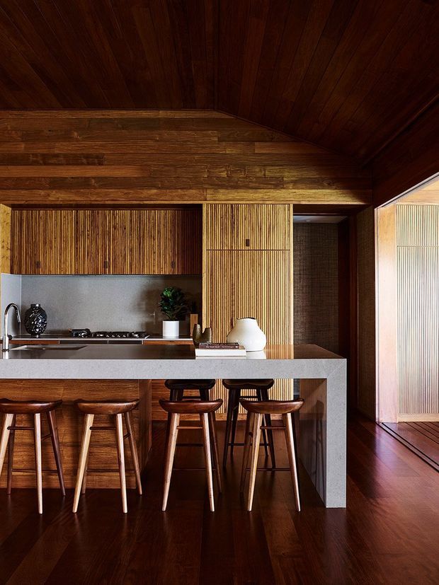 A richly timber-lined kitchen with a vaulted timber ceiling, dark wood cabinetry and flooring, and a long light-grey stone island with integrated seating. Vertical ribbed timber joinery creates a textured feature wall, while warm pendant-free lighting and carefully styled ceramics give the space a refined, intimate atmosphere. An adjacent timber-lined passage is visible to the right.