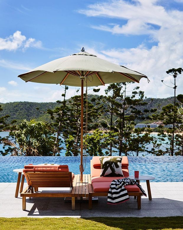 Outdoor poolside lounge area with a large cream cantilever-style umbrella, timber sun loungers, and coral-red upholstered seating arranged beside a blue swimming pool. A landscaped hillside and coastal-looking landscape are visible beyond the pool under a bright blue sky.