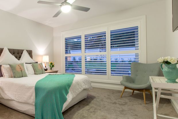 Bright, softly styled bedroom featuring a large upholstered bed with layered white and pale green bedding, a teal knitted throw, a grey upholstered accent chair, a white side table, and a large set of white plantation-shuttered windows. Neutral carpet and walls create a calm, inviting atmosphere.