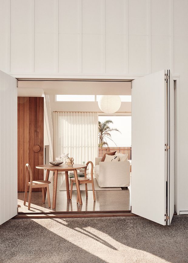 A bright, warm-toned view through wide open white paneled doors into a combined dining and living area. The room features a round timber dining table with curved wooden chairs, a tall timber cabinet, a pale upholstered armchair, sheer curtains, and a large paper pendant light. Sunlight creates strong diagonal shadows across the dark textured foreground floor, while a palm and timber privacy screen are visible through the glazing.