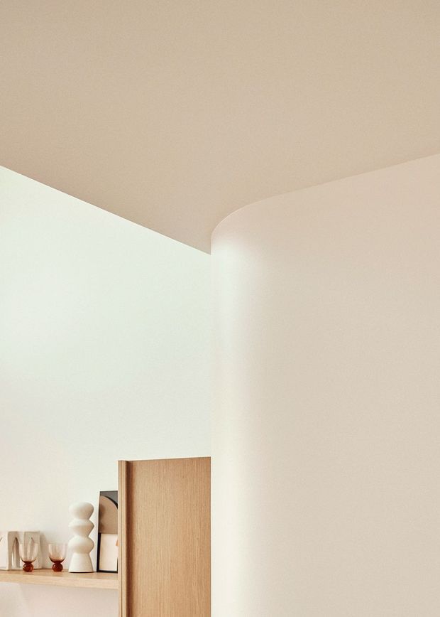 Minimalist interior detail showing a softly rounded plastered ceiling-to-wall transition in warm off-white tones. A light timber door or partition stands near the lower edge, with a small shelf displaying glassware and sculptural objects. The image emphasizes smooth curves, diffuse lighting, and restrained natural materials.