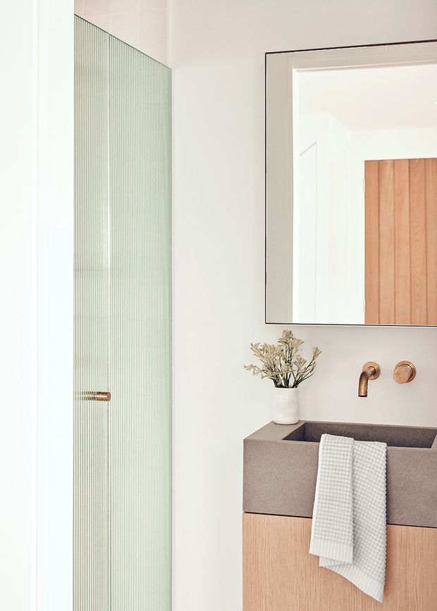 Bright contemporary bathroom featuring a wall-mounted timber vanity with a taupe stone or composite basin, brushed bronze wall-mounted tapware, a large frameless mirror, and a vertically ribbed translucent glass shower screen with a bronze handle. The palette is warm and neutral, with pale walls, light timber cabinetry, and subtle decorative dried foliage.