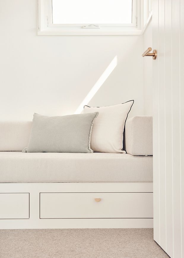 Bright, minimalist built-in window seat with upholstered bench cushions and pale textured pillows, set above integrated drawers. White vertical wall panelling, a small white-framed window, and a bronze lever door handle create a clean, soft-toned interior.