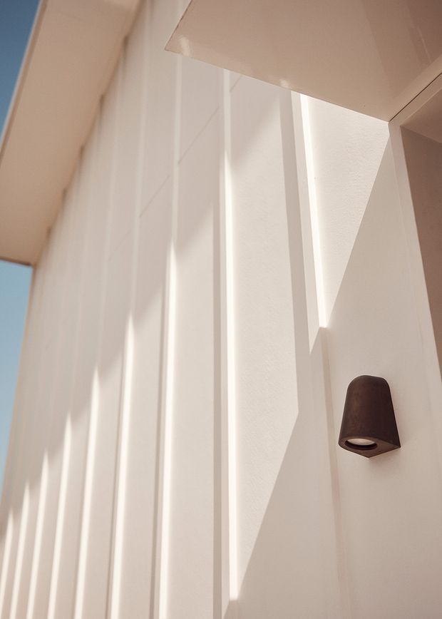 Close-up architectural detail of a white, textured exterior wall with deep vertical fins or pilasters creating strong geometric shadows. A dark bronze or brown wall-mounted light fixture provides a contrasting accent, while narrow glimpses of blue sky appear at the edges. The image has a minimalist, sunlit modern aesthetic.